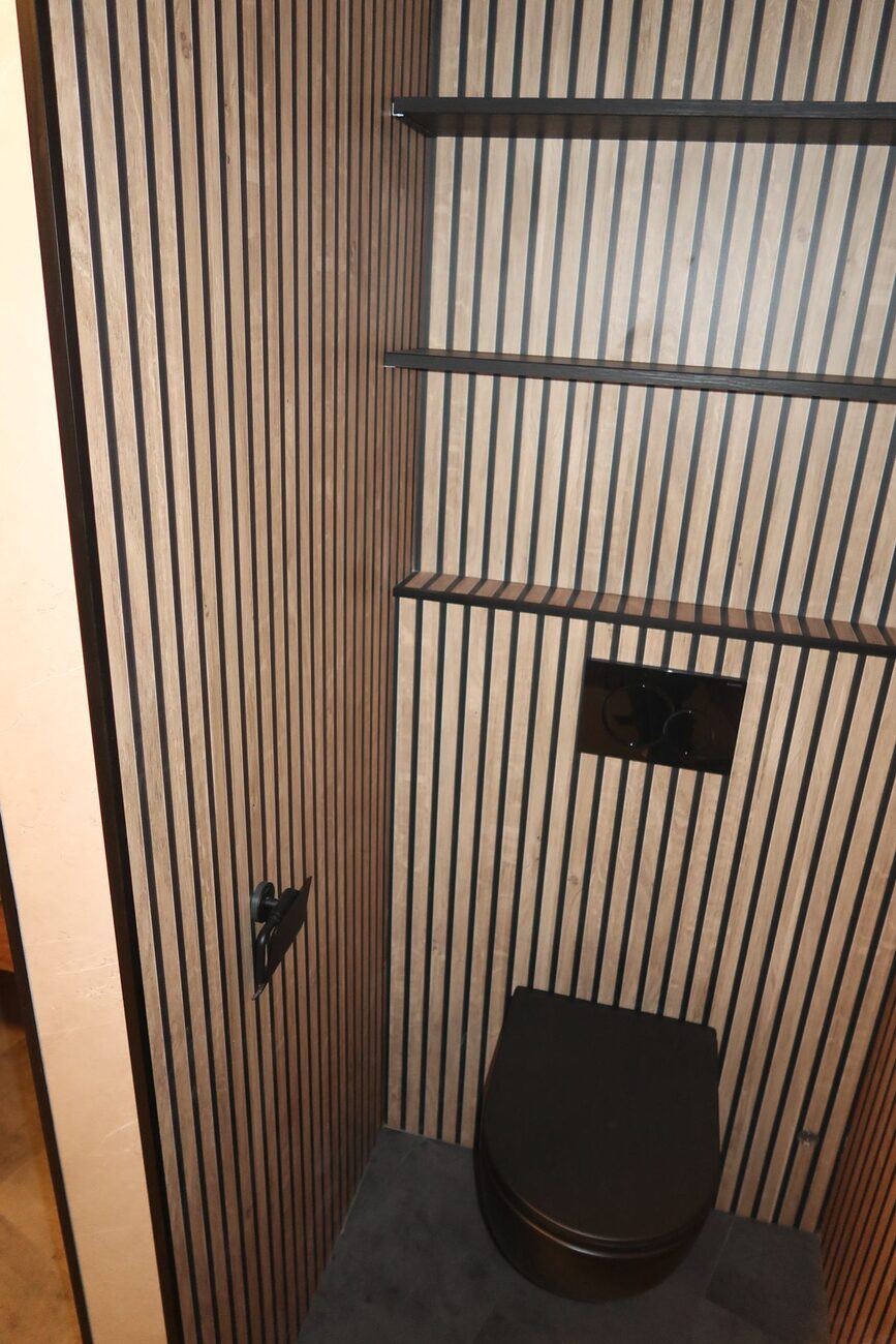 Toiletruimte met houten lattenwanden, zwarte toiletpot en planken.