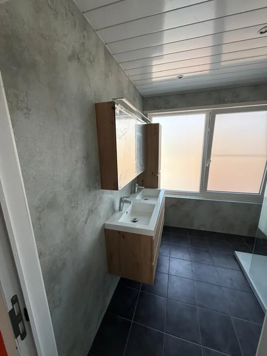 Badkamer met grijze betonlook wandpanelen, houten zwevend wastafelmeubel met spiegelkast en donkere vloertegels