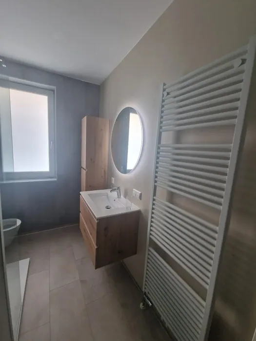 Badkamer met eiken wastafelmeubel, ronde spiegel met verlichting en grote witte handdoekradiator