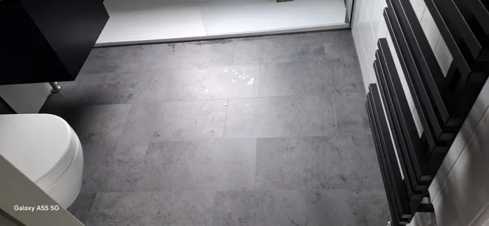 Grijze tegelvloer in betonlook met zwarte designradiator, hangtoilet en douchebak in beeld