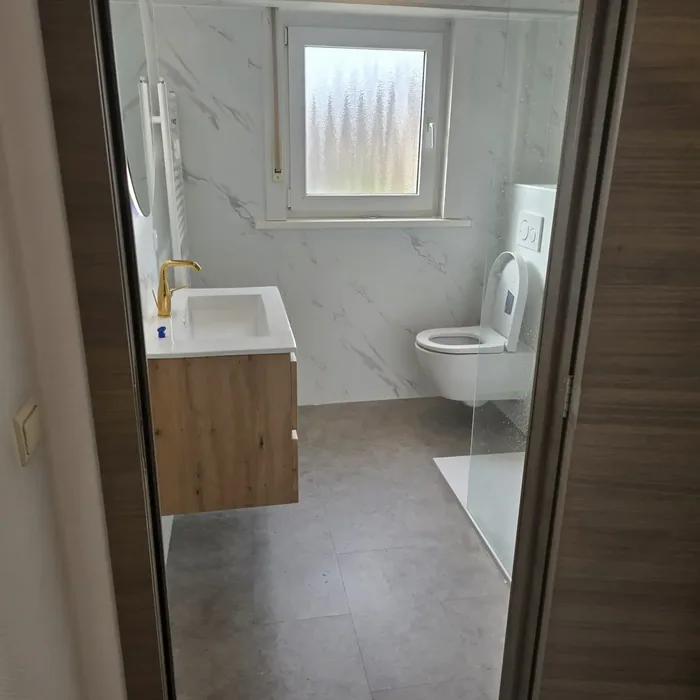 Doorkijk naar badkamer met marmerlook wandpanelen, eiken wastafelmeubel met gouden kraan en hangtoilet