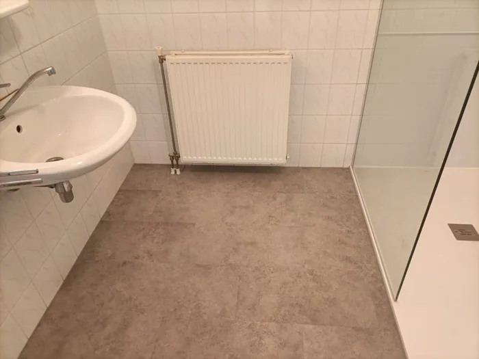 Badkamer met nieuwe steenlook vloer, wastafel, radiator en douchebak met glazen wand