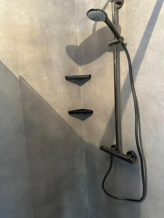 Zwarte regendoucheset met thermostaatkraan, handdouche en twee zwarte hoekplankjes tegen grijze wandpanelen