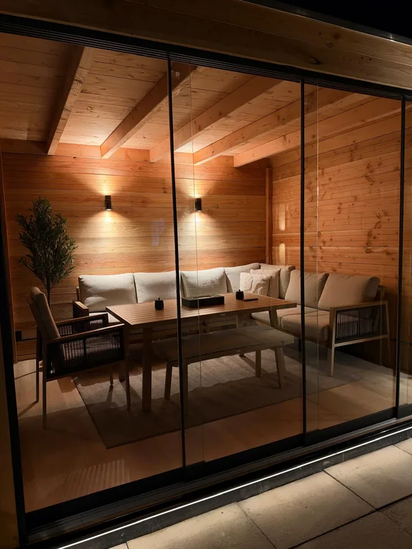 Interieur van een houten tuinkamer met balkenplafond, horizontale houten wandbetimmering, twee wandlampen en een loungeset