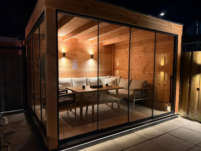 Houten tuinkamer met glazen schuifwanden, schuin van voren gezien, met houten balkenplafond, wandlampen en ledverlichting langs de onderkant