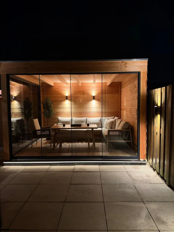 Houten veranda met glazen schuifwanden in de avond, vooraanzicht met loungebank, eettafel en een ledstrip langs de vloer