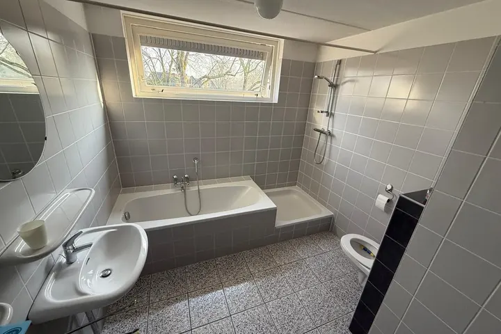 Oude badkamer met ligbad, wastafel en staand toilet, grijze tegels en gespikkelde vloer