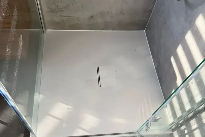 Witte douchebak met lijnafvoer en glazen wanden, betonlook wand en grijze vloer in zonlicht