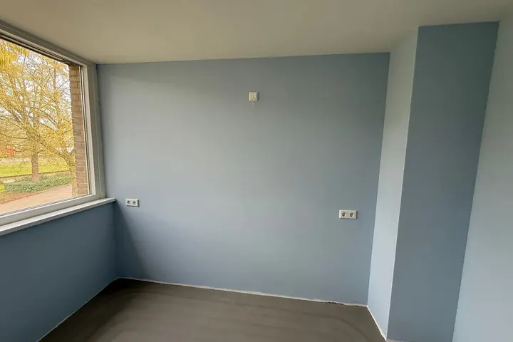 Lege kamer met blauwgrijze muren, raam met tuinzicht en kale dekvloer