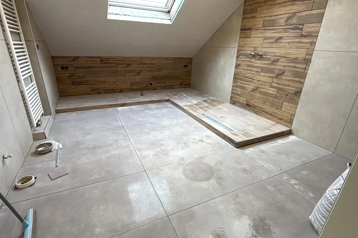 Zolderbadkamer in afbouw met grote grijze tegels, houtlook wand, dakraam en handdoekradiator