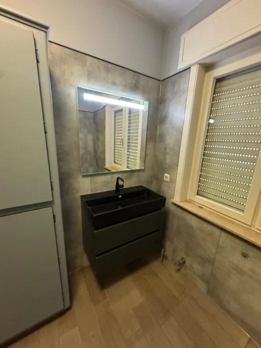 Badkamer met grijze betonlook wandpanelen, zwart wastafelmeubel, LED-spiegel, hoge kast en houtlook vloer