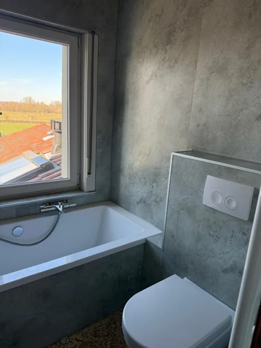 Ligbad onder het raam, wanden in grijze betonlook panelen en hangtoilet met bedieningsplaat